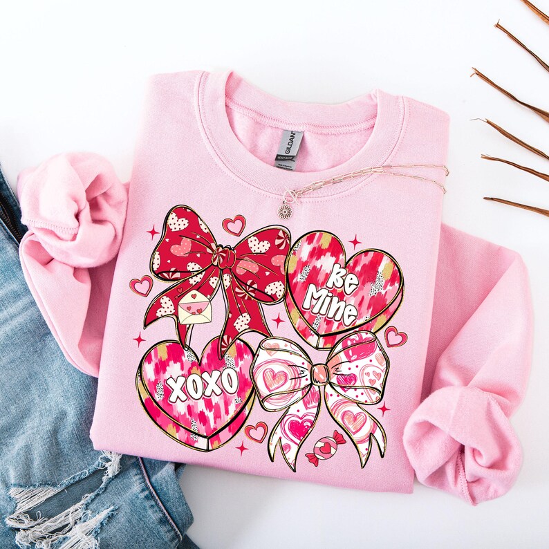 Puede incluir: Una sudadera rosa claro con un dise&ntilde;o de San Valent&iacute;n. El dise&ntilde;o incluye lazos rojos, caramelos en forma de coraz&oacute;n con las frases "Be Mine" y "XOXO", y otros gr&aacute;ficos de corazones y caramelos. Un delicado collar est&aacute; sobre la sudadera.