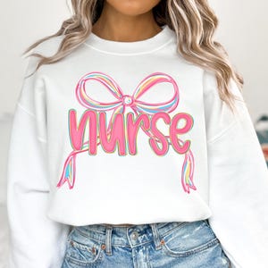 Pode incluir: Camisola branca com a palavra "Nurse" em rosa, com um design de la&ccedil;o colorido por cima. O la&ccedil;o tem detalhes em rosa pastel, amarelo e azul. Um estilo de camisola casual e confort&aacute;vel.