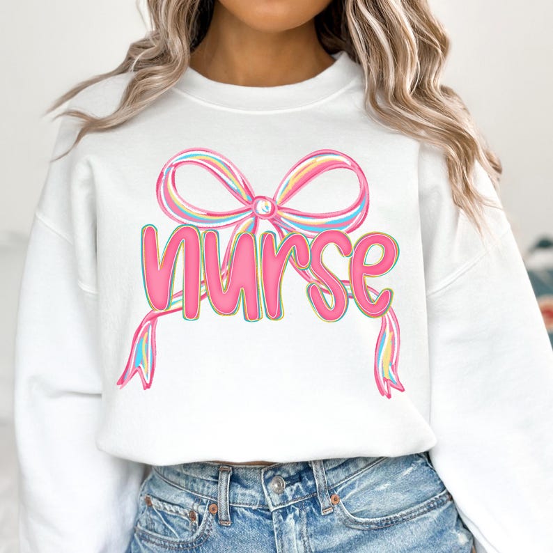 Pode incluir: Camisola branca com a palavra "Nurse" em rosa, com um design de la&ccedil;o colorido por cima. O la&ccedil;o tem detalhes em rosa pastel, amarelo e azul. Um estilo de camisola casual e confort&aacute;vel.
