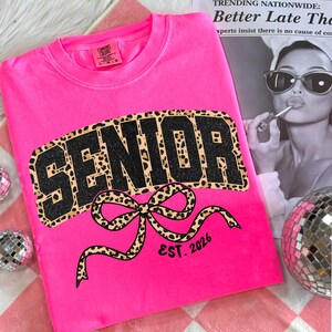 May include: A bright pink sweatshirt with the word "SENIOR" in large, black glitter letters with a leopard print border. A leopard print bow is below the word, with "EST. 2026" printed below the bow. The sweatshirt is a Comfort Colors brand.