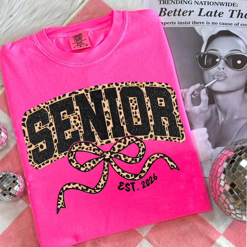 May include: A bright pink sweatshirt with the word "SENIOR" in large, black glitter letters with a leopard print border. A leopard print bow is below the word, with "EST. 2026" printed below the bow. The sweatshirt is a Comfort Colors brand.