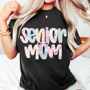 Puede incluir: Camiseta negra con la frase "Senior Mom" en un dise&ntilde;o de patchwork colorido. Las letras est&aacute;n rellenas de telas florales y estampadas en tonos rosa, azul y blanco, con un lazo rosa. Una prenda superior casual y c&oacute;moda.