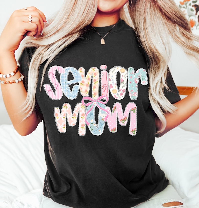 Puede incluir: Camiseta negra con la frase "Senior Mom" en un dise&ntilde;o de patchwork colorido. Las letras est&aacute;n rellenas de telas florales y estampadas en tonos rosa, azul y blanco, con un lazo rosa. Una prenda superior casual y c&oacute;moda.