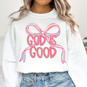 May include: White sweatshirt with a colorful bow design and the words "GOD IS GOOD" in pink. The bow has pink, yellow, and blue accents. The sweatshirt is a casual, comfortable style.