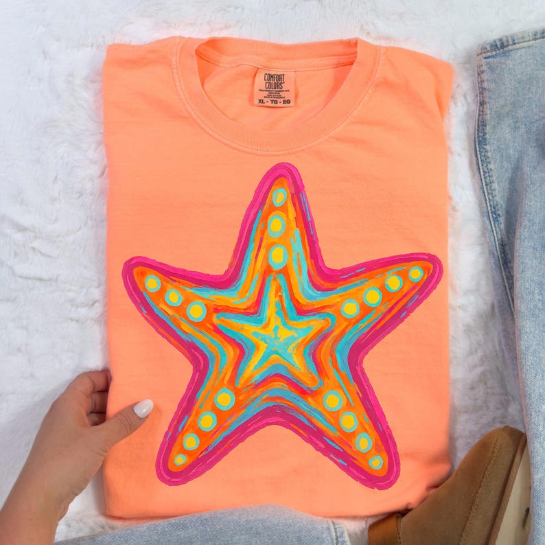 May include: Coral-colored t-shirt featuring a vibrant starfish design. The starfish is outlined in pink and filled with shades of blue, yellow, and orange. The shirt has a crew neck and short sleeves. The tag reads XL-TG-EG.