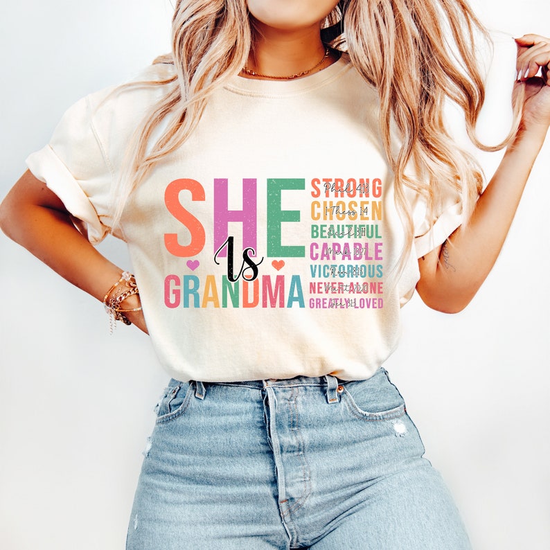 May include: A white t-shirt with the text "She is Grandma" in colorful letters. The text is stacked vertically with the word "Grandma" at the bottom. The words "Strong, Chosen, Beautiful, Capable, Victorious, Never Alone, Greatly Loved" are stacked vertically to the right of the word "She".