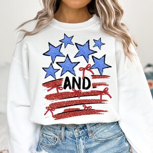 Pode incluir: Camisola branca com um gr&aacute;fico vermelho, branco e azul de uma bandeira americana estilizada com estrelas e la&ccedil;os vermelhos. O texto "AND" est&aacute; impresso em preto na bandeira.