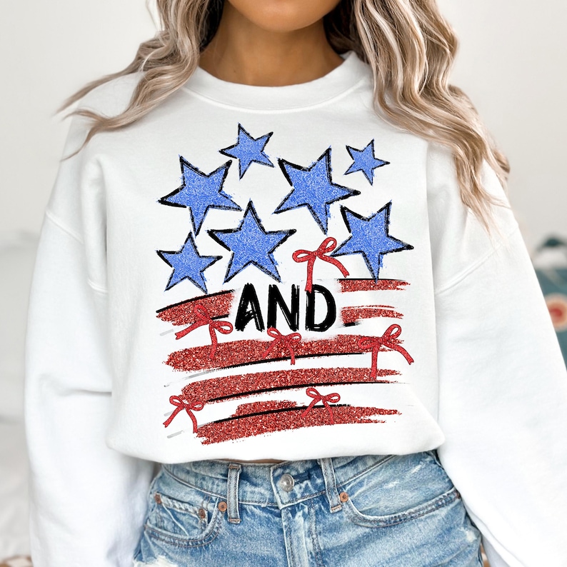 Pode incluir: Camisola branca com um gr&aacute;fico vermelho, branco e azul de uma bandeira americana estilizada com estrelas e la&ccedil;os vermelhos. O texto "AND" est&aacute; impresso em preto na bandeira.