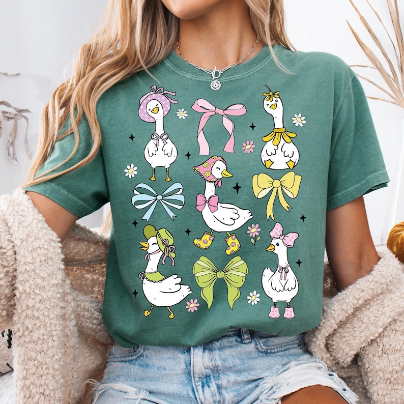 Puede incluir: Una camiseta verde con un estampado gr&aacute;fico de varios gansos de dibujos animados que llevan sombreros y sujetan lazos. Los gansos est&aacute;n en varias poses y tienen sombreros y lazos de diferentes colores.