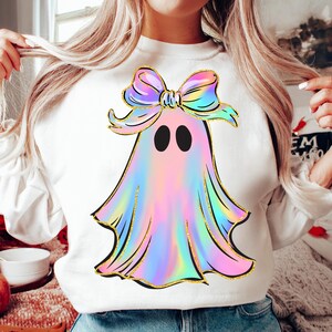 May include: White sweatshirt featuring a colorful ghost design. The ghost has a rainbow-colored body, black eyes, and a bow. The design is outlined in gold glitter. A fun and festive top for Halloween.