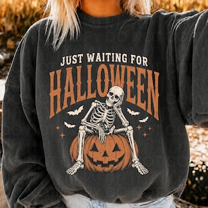 May include: A charcoal gray long-sleeve sweatshirt featuring a graphic print of a skeleton sitting on a jack-o'-lantern pumpkin. The text reads JUST WAITING FOR HALLOWEEN in a distressed font. Small white bats and four-pointed stars surround the skeleton.