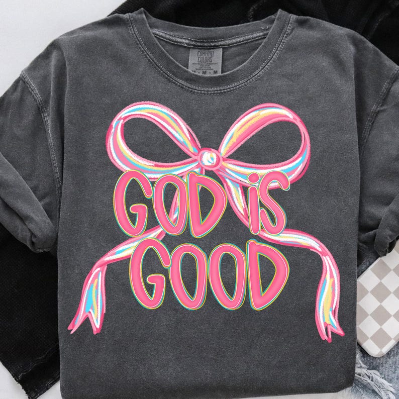 May include: Dark gray long-sleeve shirt with a colorful bow graphic and the text "GOD IS GOOD" in pink. The bow features pastel rainbow colors. The shirt is a comfortable, casual style.