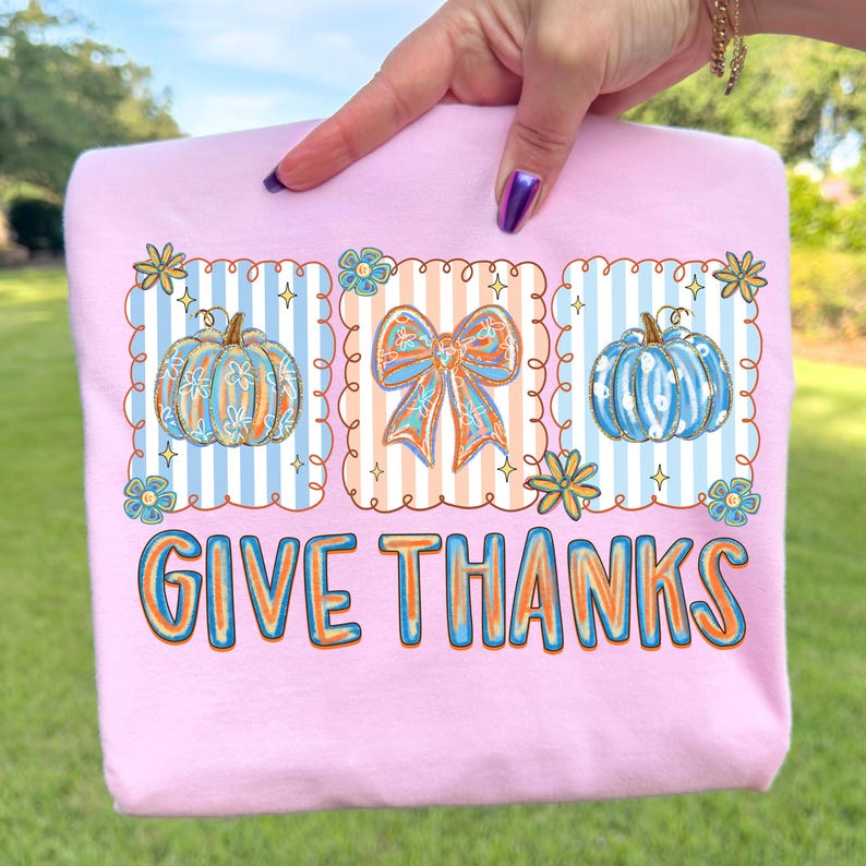 May include: A light pink sweatshirt with a fall-themed graphic. The graphic includes two pumpkins, a bow, and the words "GIVE THANKS" in orange and blue. The pumpkins and bow are decorated with blue and orange accents.