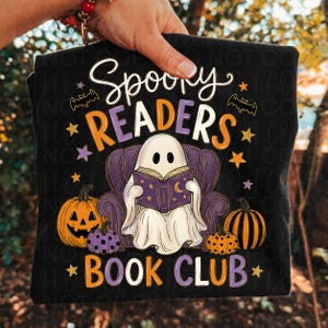 May include: A black t-shirt featuring a graphic of a white ghost sitting in a purple armchair reading a purple book. The text reads Spooky Readers Book Club. The design includes orange and purple pumpkins, small stars, and bats in orange and purple.