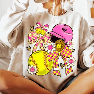 May include: Cream-colored sweatshirt featuring a softball-themed graphic. The design includes a pink helmet, yellow softball, and a brown glove, accented with pink and yellow bows, hearts, and floral elements. The overall theme is softball.