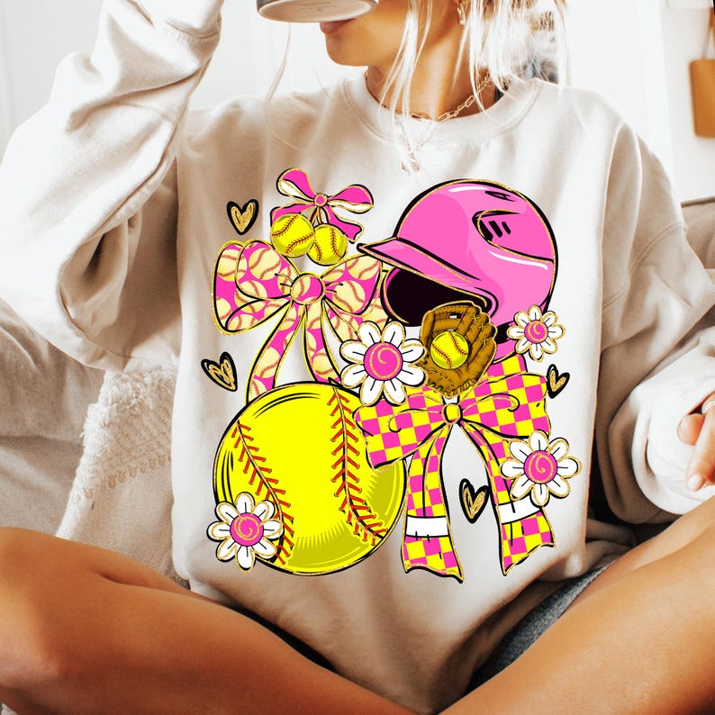May include: Cream-colored sweatshirt featuring a softball-themed graphic. The design includes a pink helmet, yellow softball, and a brown glove, accented with pink and yellow bows, hearts, and floral elements. The overall theme is softball.