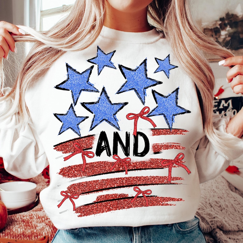 Pode incluir: Camisola branca com um design de bandeira americana vermelha, branca e azul. A bandeira &eacute; feita de estrelas e listras com la&ccedil;os vermelhos. A palavra "AND" est&aacute; escrita em letras pretas na bandeira.
