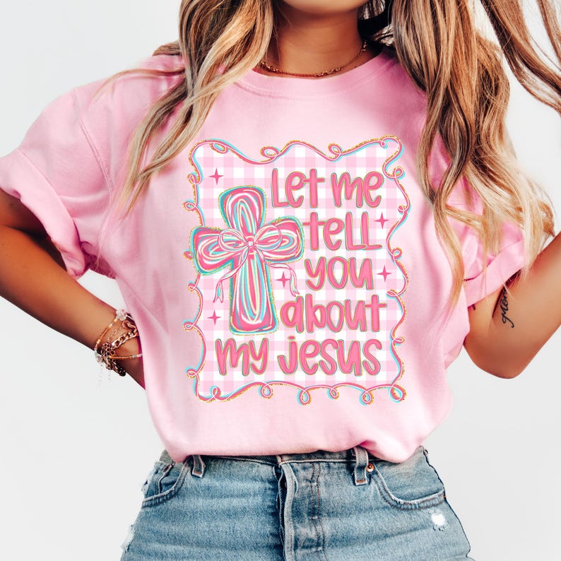 May include: Pink t-shirt with a graphic featuring a cross with a bow and the text "Let me tell you about my Jesus." The cross is in shades of pink, blue, and green, set against a gingham background.
