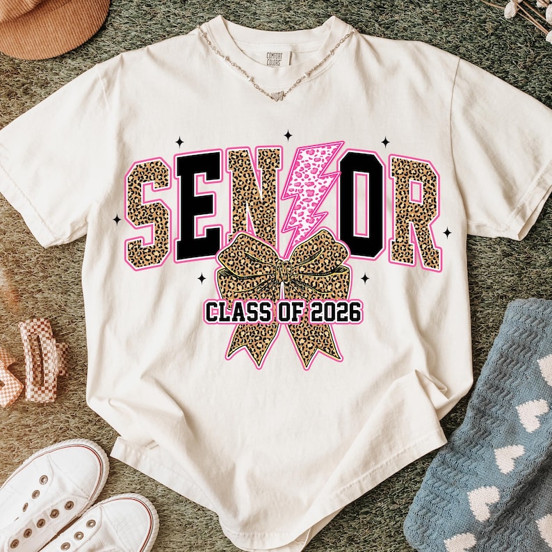 Pu&ograve; includere: T-shirt color crema con la scritta "SENIOR" in nero e stampa leopardata, un fulmine rosa e un fiocco leopardato. Il testo "CLASS OF 2026" &egrave; sotto il fiocco. La maglietta &egrave; circondata da accessori.
