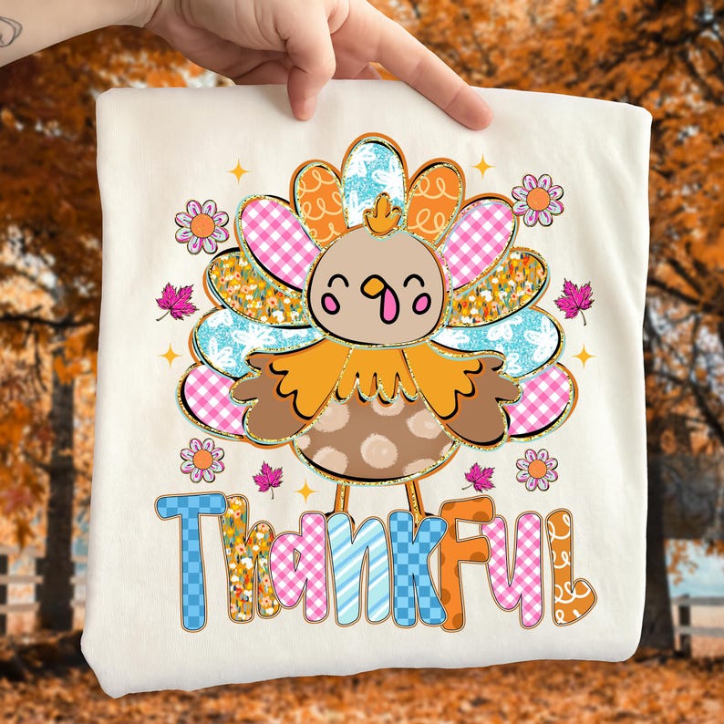 May include: A cream-colored t-shirt featuring a colorful turkey design with the word "Thankful" in a playful font. The turkey has a brown body, orange feathers, and a cheerful expression. Floral and leaf accents surround the turkey.