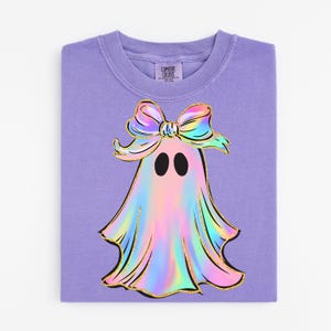 May include: A lavender Comfort Colors crewneck sweatshirt featuring a colorful ghost design. The ghost has a holographic appearance with a gold outline and a bow. The ghost has two black oval eyes. The sweatshirt is folded.