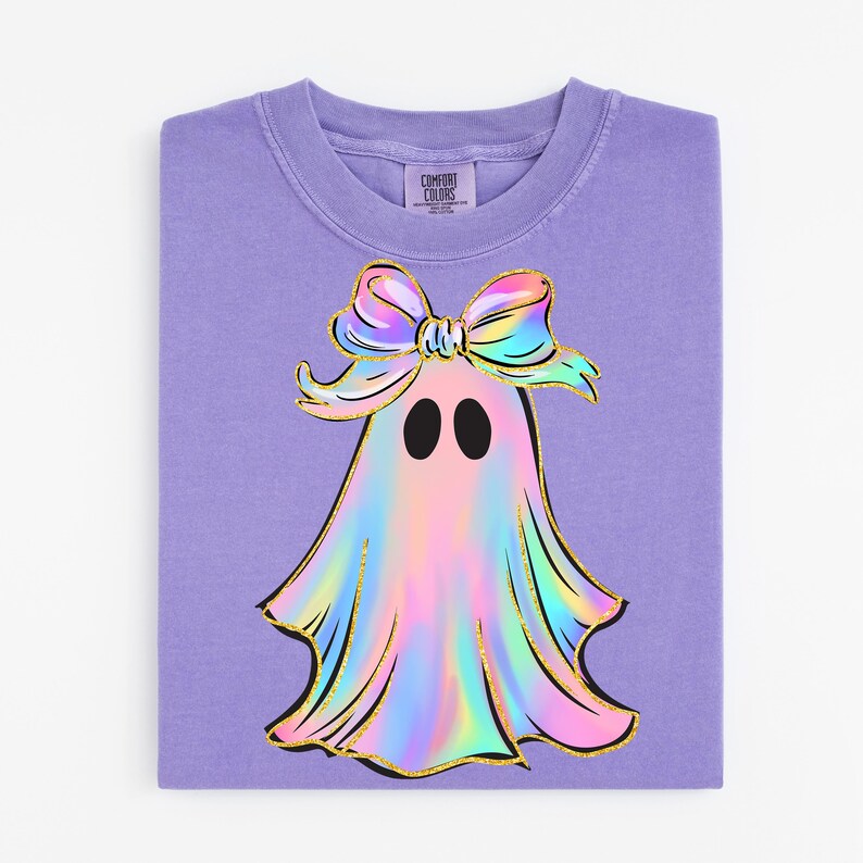 May include: A lavender Comfort Colors crewneck sweatshirt featuring a colorful ghost design. The ghost has a holographic appearance with a gold outline and a bow. The ghost has two black oval eyes. The sweatshirt is folded.