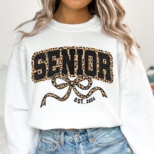 May include: White sweatshirt with a black and leopard print "SENIOR" design. A leopard print bow with "EST. 2026" is below the word "SENIOR". The sweatshirt is a crew neck style.