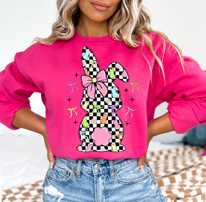 May include: A pink sweatshirt with a checkered bunny rabbit design. The bunny is made up of pastel-colored jelly beans and has a pink bow. The design is surrounded by small white flowers and pastel-colored bows.