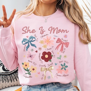 K&ouml;nnte beinhalten: Rosafarbener Pullover mit dem Text "She Is Mom" in rosa Schrift. Der Pullover hat ein florales Design mit Schleifen und Schmetterlingen. Das Design enth&auml;lt auch Bibelverse und Daten.
