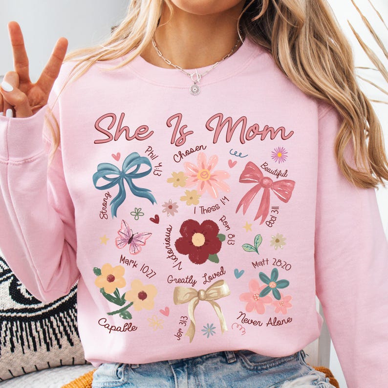 K&ouml;nnte beinhalten: Rosafarbener Pullover mit dem Text "She Is Mom" in rosa Schrift. Der Pullover hat ein florales Design mit Schleifen und Schmetterlingen. Das Design enth&auml;lt auch Bibelverse und Daten.