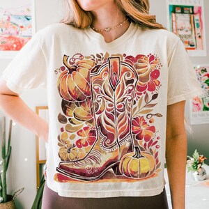 Op de afbeelding: Cr&egrave;mekleurig T-shirt met een kleurrijke afbeelding van een cowboylaars, pompoenen en bloemelementen in oranje, rood en bruin. Het ontwerp heeft een herfst- of oogstthema.