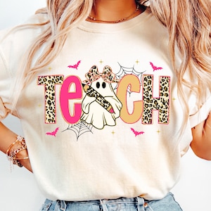 May include: Cream-colored sweatshirt with the word "TEACH" in pink and tan, with leopard print accents. A ghost holding a pencil is in the center. Pink bats and spiderwebs decorate the design, perfect for teachers during Halloween.