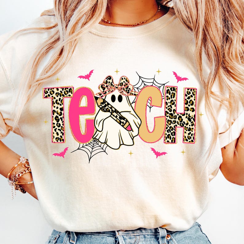 May include: Cream-colored sweatshirt with the word "TEACH" in pink and tan, with leopard print accents. A ghost holding a pencil is in the center. Pink bats and spiderwebs decorate the design, perfect for teachers during Halloween.
