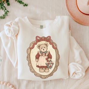 Op de afbeelding: Cr&egrave;mekleurige sweatshirt met een vintage-stijl illustratie van een teddybeer. De beer draagt een rood-witte jurk en houdt een kleine quilt vast. De illustratie is omlijst door een decoratief ovaal met een strik aan de bovenkant.