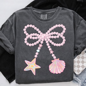 May include: A grey t-shirt featuring a pink pearl bow, two starfish, and a seashell. The bow is made of pearls and has a pink centre. The starfish are yellow, orange, and pink. The seashell is pink and white.