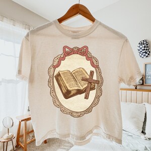 May include: Cream-colored t-shirt featuring a central graphic of an open book and a wooden cross within a decorative oval frame. The frame is adorned with a pink bow at the top. The shirt is hanging on a wooden hanger.