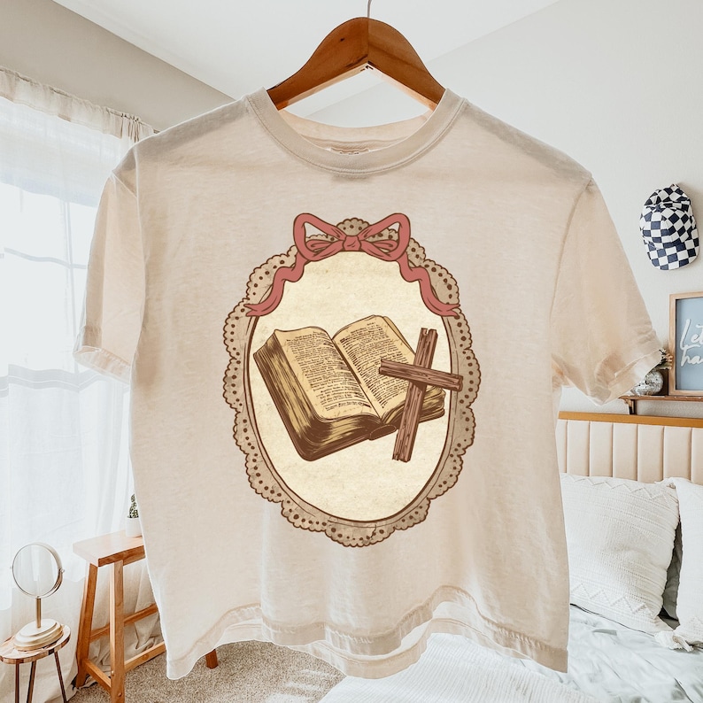 May include: Cream-colored t-shirt featuring a central graphic of an open book and a wooden cross within a decorative oval frame. The frame is adorned with a pink bow at the top. The shirt is hanging on a wooden hanger.
