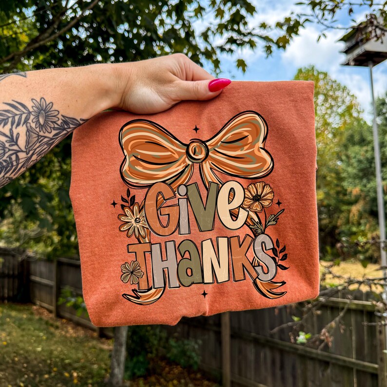 May include: An orange-colored t-shirt with the words "GIVE THANKS" in a decorative font. The design includes a large bow, flowers, and leaves in shades of brown, beige, and green. The shirt is held up by a hand with a floral tattoo.