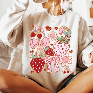 Op de afbeelding: Een wit sweatshirt met een kleurrijke grafische print van aardbeien, strikken en bloemen. Het ontwerp heeft rode, roze, groene en gouden accenten.