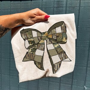 May include: A beige t-shirt featuring a large bow design. The bow is composed of various camouflage patterns and white and green checkered sections. The shirt is held up by a hand with red nail polish against a blue background.