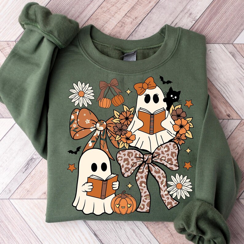Op de afbeelding: Olijfgroene sweatshirt met een Halloween-thema ontwerp. Het ontwerp bevat twee spookfiguren die boeken lezen, omringd door pompoenen, strikken, bloemen, sterren en een zwarte kat. De strikken hebben bloemen- en luipaardprints.