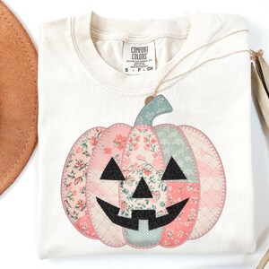 May include: Cream-colored t-shirt featuring a patchwork pumpkin design. The pumpkin is composed of pink, floral, and patterned fabrics with a black face. A gold necklace hangs over the shirt. The shirt has a Comfort Colors tag.