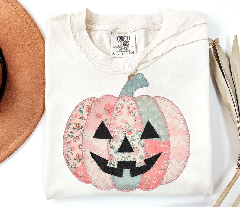 May include: Cream-colored t-shirt featuring a patchwork pumpkin design. The pumpkin is composed of pink, floral, and patterned fabrics with a black face. A gold necklace hangs over the shirt. The shirt has a Comfort Colors tag.