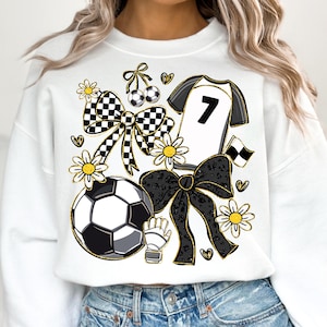 May include: White sweatshirt with a soccer-themed design. Features a soccer ball, jersey with the number 7, bows, flowers, and a soccer glove. The design incorporates black, white, and gold accents, creating a playful and sporty aesthetic.