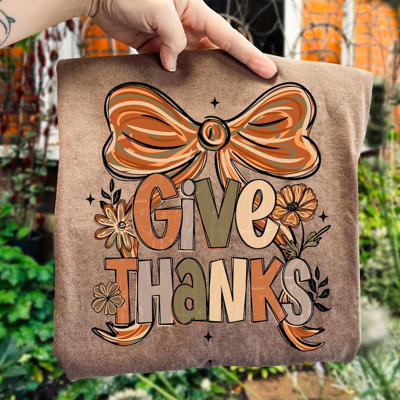 May include: A brown t-shirt with the words "Give Thanks" in a decorative font. The design includes a large bow, flowers, and leaves in shades of orange, brown, and green. The shirt is being held up by a hand.
