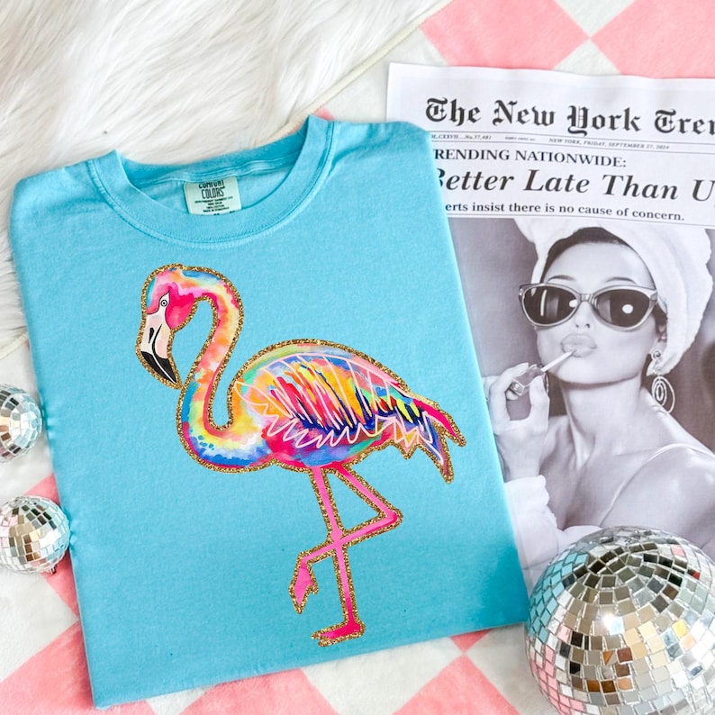 May include: A light blue t-shirt featuring a colorful flamingo design with gold glitter accents. The flamingo has a rainbow-colored body and pink legs. The shirt is on a pink and white checkered surface, with a newspaper clipping and disco balls.