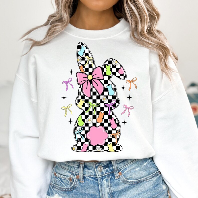 May include: White sweatshirt with a checkered bunny silhouette design. The bunny is decorated with pastel-colored bows, flowers, and jelly beans.