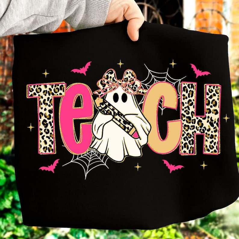 May include: Black t-shirt with the word "TEACH" in a leopard print and pink font. A ghost wearing a leopard print bow and holding a pencil is in the center. Pink bats, spiderwebs, and stars decorate the design.