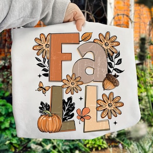 May include: White tote bag featuring the word "FALL" in brown, tan, and orange block letters. The design includes fall-themed illustrations such as flowers, a pumpkin, an acorn, a leaf, and a butterfly. The bag is held by a person.