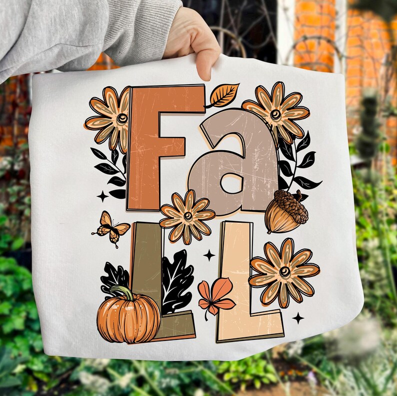 May include: White tote bag featuring the word "FALL" in brown, tan, and orange block letters. The design includes fall-themed illustrations such as flowers, a pumpkin, an acorn, a leaf, and a butterfly. The bag is held by a person.